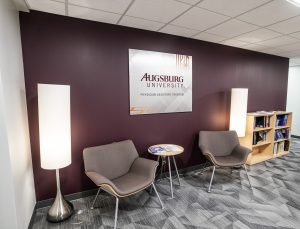 Reception area featuring two lobby chairs against a maroon wall with Augsburg University metal artwork. Chairs are framed by floor lamps 